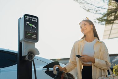 Alışveriş çantası tutan genç bir kadın, alışveriş merkezinin otoparkındaki şarj istasyonundan EV 'nin aküsünü şarj etmek için akıllı telefon kullanıyor. Modern kadın eco arabasıyla alışverişe gider. Yararlı