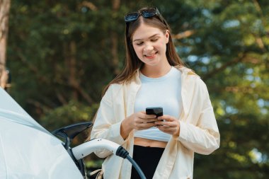 Genç bir kadın EV otomobil şarj istasyonu Green City Park 'ta elektrik ödemek için akıllı telefonu kullanıyor. Modern çevre ve EV araçlı sürdürülebilir şehir yaşam tarzı. Yararlı