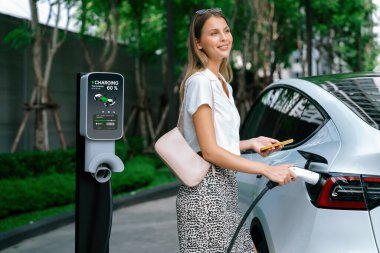 Genç bir kadın yazın yeşil sürdürülebilir şehir bahçesinde EV elektrikli arabasıyla seyahat ediyor. Kentsel sürdürülebilirlik yaşam tarzı yeşil temiz yeniden şarj edilebilir enerji elektrikli BEV araç iç organları