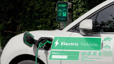EV şarj kablosuna elektrik kablosu takılı. Çevre yeşil parkı ve yeşillik arka planında akıllı dijital batarya durum hologramını gösteriyor. Enerji sürdürülebilirliği teknolojik gelişim.