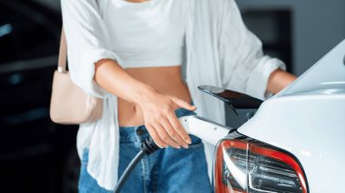 Genç bir kadın EV elektrikli arabasıyla alışveriş merkezinin otoparkına gidiyor şehir merkezinde elektrik iç organlarının yeşil temiz şarj edilebilir enerji ile sürdürülebilirlik yaşam tarzını gösteriyor.