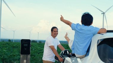 İlerici mutlu aile kavramı elektrikli araçla rüzgar çiftliğinde eğleniyor. Rüzgar türbini jeneratöründen gelen temiz yenilenebilir enerji ile şarj edilen elektrikli araç..