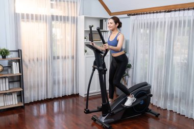 Enerjik ve güçlü atletik Asyalı kadın evde eliptik koşu makinesiyle koşuyor. Formda fiziğin takibi ve ev çalışması ve eğitimle sağlıklı yaşam tarzına bağlılık. Kuvvetli