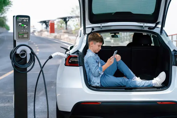 小男孩坐在汽车行李箱上 用智能手机充电 同时从电动车充电站给环保车充电 电动汽车汽车作为可持续能源概念下的代用车辆的公路旅行 永远不变 — 图库照片