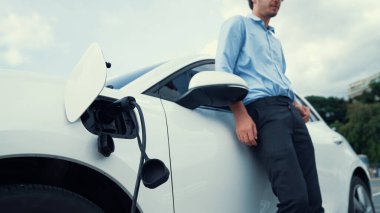 Takım elbiseli, hırslı bir işadamı elektrik kablosu ve yenilenebilir enerji enerjili bir elektrikli araçla şarj istasyonunda dururken elektrikli aracına yaslanıyor..