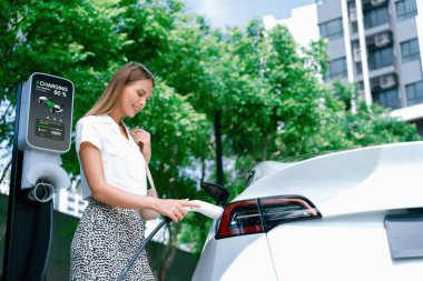 Genç bir kadın yazın yeşil sürdürülebilir şehir bahçesinde EV elektrikli arabasıyla seyahat ediyor. Kentsel sürdürülebilirlik yaşam tarzı yeşil temiz yeniden şarj edilebilir enerji elektrikli BEV araç iç organları