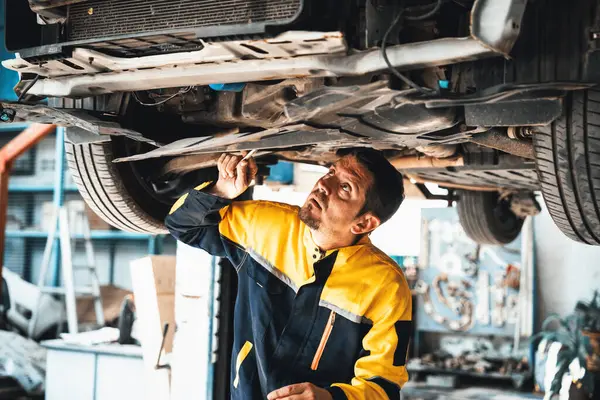 Vehicle mechanic conduct car inspection from beneath lifted vehicle. Automotive service technician in uniform carefully diagnosing and checking cars axles and undercarriage components. Oxus