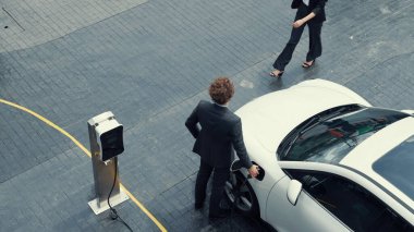 İlerici iş adamı ve iş kadını şehir merkezinde dolaşmadan önce şarj istasyonuna bağlı elektrikli arabaya yaslanıyor. Çevre dostu şarj edilebilir araba temiz enerjiyle çalışıyor..