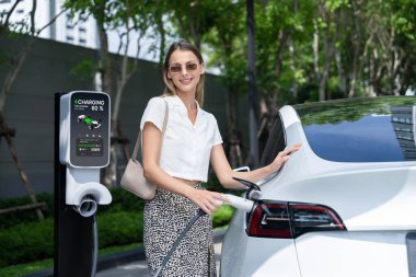 Genç bir kadın yazın yeşil sürdürülebilir şehir bahçesinde EV elektrikli arabasıyla seyahat ediyor. Kentsel sürdürülebilirlik yaşam tarzı yeşil temiz yeniden şarj edilebilir enerji elektrikli BEV araç iç organları