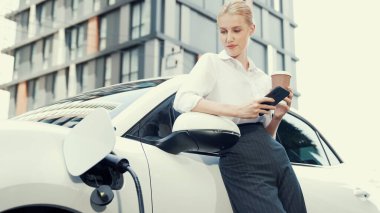 Şehir merkezinde dolaşmadan önce elektrikli araba ve şarj istasyonuna yaslanan ilerici iş kadını. Çevre dostu şarj edilebilir EV araba sürdürülebilir ve temiz enerjiyle çalışıyor.
