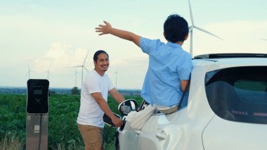 İlerici mutlu aile kavramı elektrikli araçla rüzgar çiftliğinde eğleniyor. Rüzgar türbini jeneratöründen gelen temiz yenilenebilir enerji ile şarj edilen elektrikli araç..