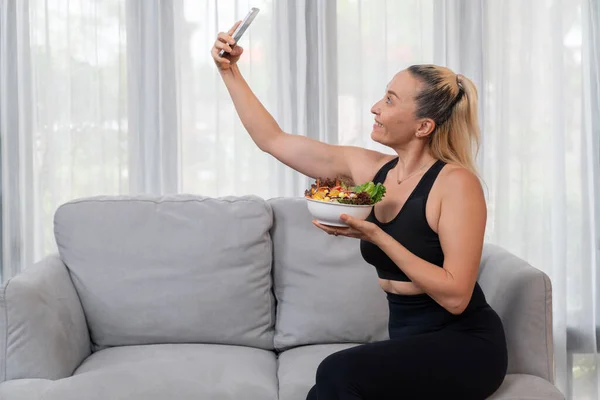 Healthy senior woman in sportswear taking selfie with smartphone, holding a bowl of fruit and vegetable. Vegan lifestyle and healthy cuisine nutrition for fitness body physique concept. Clout