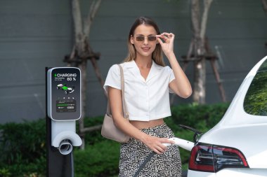 Genç bir kadın yazın yeşil sürdürülebilir şehir bahçesinde EV elektrikli arabasıyla seyahat ediyor. Kentsel sürdürülebilirlik yaşam tarzı yeşil temiz yeniden şarj edilebilir enerji elektrikli BEV araç iç organları