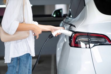 Genç bir kadın EV elektrikli arabasıyla alışveriş merkezinin otoparkına gidiyor şehir merkezinde elektrik iç organlarının yeşil temiz şarj edilebilir enerji ile sürdürülebilirlik yaşam tarzını gösteriyor.