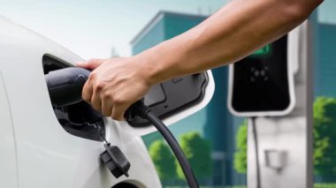 Adam elektrikli araca akıllı EV şarj cihazı takıyor ve arka planda yeşil şehir parkı olan EV şarj istasyonundan şarj oluyor. ESG şehrinde gelecekteki sürdürülebilir güç ve EV araba kullanımı. İnceleyin