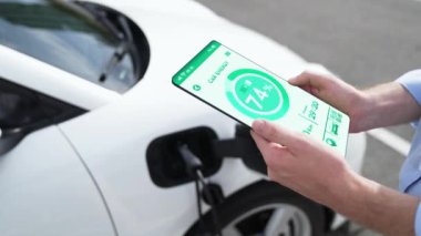 Akıllı telefon ekran bataryasını elinde tutan iş adamı akıllı EV mobil uygulama ile otomobil parkındaki şarj istasyonundan elektrik şarj eden çevre dostu EV otomobil. İnceleyin