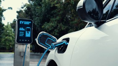 Elektrikli araba, çevre yeşil parkındaki elektrik kablosu ve yeşillik yeşili yeşillik yapraklarıyla şarj etmek için şarj istasyonuna bağlandı. Geleceğin yenilikçi her arabası ve enerji sürdürülebilirliği.