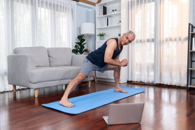 Aktif ve formda bir adam, evde egzersiz yapmadan önce ısınma ve esneme hareketleri yapıyor ve çevrimiçi fitness videosu izliyor. Emekliler için sağlıklı yaşam tarzı konsepti. Etki