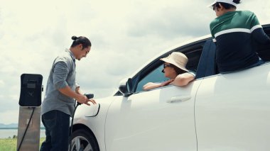 İlerici mutlu aile kavramı elektrikli araçla rüzgar çiftliğinde eğleniyor. Rüzgar türbini jeneratöründen gelen temiz yenilenebilir enerji ile şarj edilen elektrikli araç..