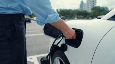 İlerici işadamı, şehir merkezinde dolaşmadan önce şarj cihazından elektrikli arabasına şarj prizi takıyor. Çevre dostu şarj edilebilir araba, sürdürülebilir ve temiz enerjiyle çalışıyor..