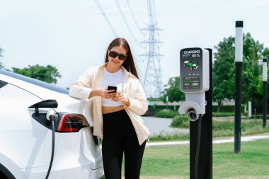 Genç bir kadın EV otomobil şarj istasyonu Green City Park 'ta elektrik ödemek için akıllı telefonu kullanıyor. Modern çevre ve EV araçlı sürdürülebilir şehir yaşam tarzı. Yararlı