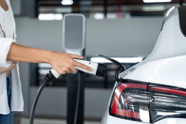 Genç bir kadın EV elektrikli arabasıyla alışveriş merkezinin otoparkına gidiyor şehir merkezinde elektrik iç organlarının yeşil temiz şarj edilebilir enerji ile sürdürülebilirlik yaşam tarzını gösteriyor.