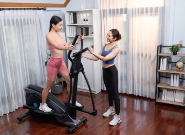 Enerjik ve güçlü atletik Asyalı kadın evde spor arkadaşı ya da antrenörü ile birlikte eliptik koşu makinesiyle koşuyor. Formda fiziğin takibi ve sağlıklı yaşam tarzına bağlılık. Kuvvetli