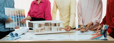 Profesyonel mimar takımının ev rengi seçimi hakkında beyin fırtınası yaparken, mimar da plan ve ev modelinin olduğu masadaki taslak belgeye yazı yazıyor. Değiştirilmiş.