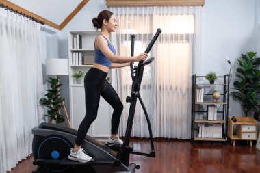 Enerjik ve güçlü atletik Asyalı kadın evde eliptik koşu makinesiyle koşuyor. Formda fiziğin takibi ve ev çalışması ve eğitimle sağlıklı yaşam tarzına bağlılık. Kuvvetli