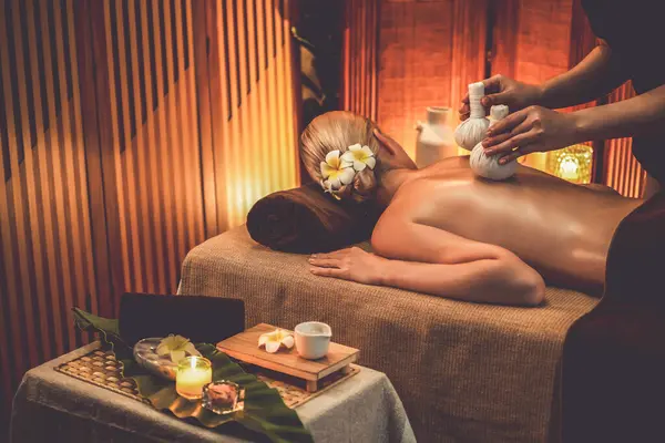 Hot herbal ball spa massage body treatment, masseur gently compresses herb bag on woman body. Tranquil and serenity of aromatherapy recreation in warm lighting of candles at spa salon. Quiescent