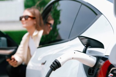 Genç bir kadın EV otomobil şarj istasyonu Green City Park 'ta elektrik ödemek için akıllı telefonu kullanıyor. Modern çevre ve EV araçlı sürdürülebilir şehir yaşam tarzı. Yararlı