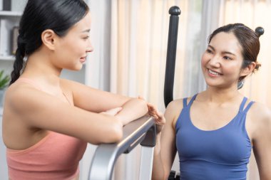 Enerjik ve güçlü atletik Asyalı kadın evde spor arkadaşı ya da antrenörü ile birlikte eliptik koşu makinesiyle koşuyor. Formda fiziğin takibi ve sağlıklı yaşam tarzına bağlılık. Kuvvetli