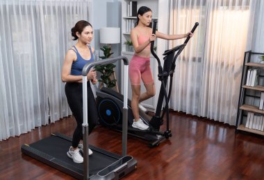 Enerjik ve güçlü atletik Asyalı kadın evde spor arkadaşı ya da antrenörü ile birlikte eliptik koşu makinesiyle koşuyor. Formda fiziğin takibi ve sağlıklı yaşam tarzına bağlılık. Kuvvetli