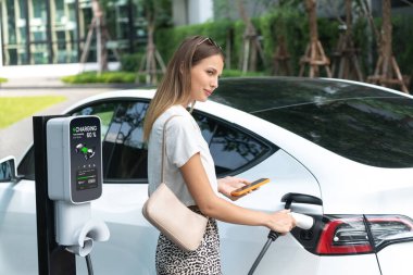Genç bir kadın yazın yeşil sürdürülebilir şehir bahçesinde EV elektrikli arabasıyla seyahat ediyor. Kentsel sürdürülebilirlik yaşam tarzı yeşil temiz yeniden şarj edilebilir enerji elektrikli BEV araç iç organları