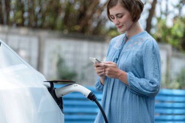 Yeşil kırsal kesimde tatilde güzel bir genç kadın elektrikli araçla şarj olurken akü durumunu kontrol ediyor. Çevre dostu seyahat zekası. Kalıcı.