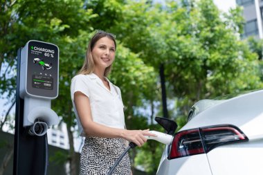 Genç bir kadın yazın yeşil sürdürülebilir şehir bahçesinde EV elektrikli arabasıyla seyahat ediyor. Kentsel sürdürülebilirlik yaşam tarzı yeşil temiz yeniden şarj edilebilir enerji elektrikli BEV araç iç organları