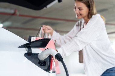 Genç bir kadın EV elektrikli arabasıyla alışveriş merkezinin otoparkına gidiyor şehir merkezinde elektrik iç organlarının yeşil temiz şarj edilebilir enerji ile sürdürülebilirlik yaşam tarzını gösteriyor.