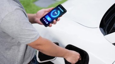 Modern yaşam tarzı adamı EV şarj cihazını ev şarj istasyonuna yerleştirir ve akıllı telefondan EV batarya durumunu etkinleştirir. Gelecekçi enerji sürdürülebilirliği ve elektrikli araba. İnceleyin