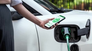 Akıllı telefon akü durumu arayüzünü elinde tutan iş kadını akıllı EV mobil uygulama ile otomobil parkındaki şarj istasyonundan elektrik şarj eden çevre dostu EV araba. İnceleyin