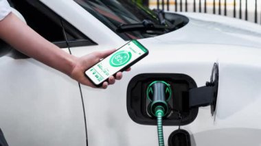 Akıllı telefon akü durumu arayüzünü elinde tutan iş kadını akıllı EV mobil uygulama ile otomobil parkındaki şarj istasyonundan elektrik şarj eden çevre dostu EV araba. İnceleyin