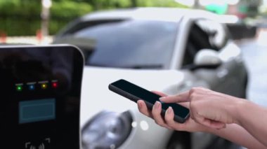 Modern iş kadını akıllı telefonun 3D hologramından batarya durumunu kontrol ederken EV arabasının şarj olan enerjisini kontrol eder. Gelecekteki EV otomotiv teknolojisinin yenilikçi teknolojik gelişimi. İnceleyin