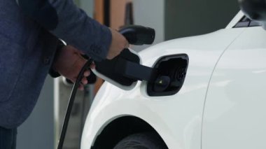 Adam garajdaki şarj istasyonundan elektrikli arabasını EV şarj cihazıyla şarj ediyor. EV arabaların teknolojik gelişimi ve gelecekteki sürdürülebilir enerji için ev enerjisi altyapısı. İnceleyin