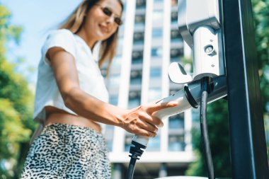 Genç bir kadın yazın yeşil sürdürülebilir şehir bahçesinde EV elektrikli arabasıyla seyahat ediyor. Kentsel sürdürülebilirlik yaşam tarzı yeşil temiz yeniden şarj edilebilir enerji elektrikli BEV araç iç organları