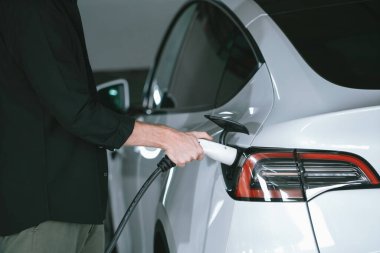 Genç adam EV elektrikli arabasıyla alışveriş merkezinin otoparkına gidiyor şehir merkezindeki şarj oluyor yeşil, temiz, yeniden şarj edilebilir elektrikli araç iç organları ile kentsel sürdürülebilirlik yaşam tarzını gösteriyor