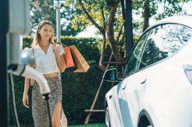 Genç bir kadın yazın yeşil sürdürülebilir şehir bahçesinde EV elektrikli arabasıyla seyahat ediyor. Kentsel sürdürülebilirlik yaşam tarzı yeşil temiz yeniden şarj edilebilir enerji elektrikli BEV araç iç organları