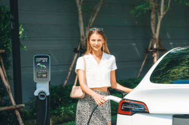 Genç bir kadın yazın yeşil sürdürülebilir şehir bahçesinde EV elektrikli arabasıyla seyahat ediyor. Kentsel sürdürülebilirlik yaşam tarzı yeşil temiz yeniden şarj edilebilir enerji elektrikli BEV araç iç organları