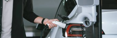 Genç adam EV elektrikli arabasıyla alışveriş merkezinin otoparkına gidiyor şehir merkezindeki şarj oluyor yeşil, temiz, yeniden şarj edilebilir elektrikli araç iç organları ile kentsel sürdürülebilirlik yaşam tarzını gösteriyor