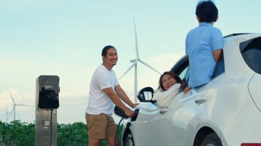 İlerici mutlu aile kavramı elektrikli araçla rüzgar çiftliğinde eğleniyor. Rüzgar türbini jeneratöründen gelen temiz yenilenebilir enerji ile şarj edilen elektrikli araç..