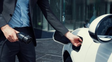 İlerici işadamı, şehir merkezinde dolaşmadan önce şarj cihazından elektrikli arabasına şarj prizi takıyor. Çevre dostu şarj edilebilir araba, sürdürülebilir ve temiz enerjiyle çalışıyor..