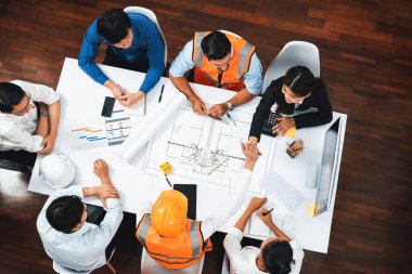 Mimari proje üzerinde birlikte çalışan, inşaat planını gözden geçiren ve toplantı masasında plan oluşturan çeşitli inşaat mühendisi ve müşteri gruplarından oluşan üst düzey bir pankart. Tedbirli